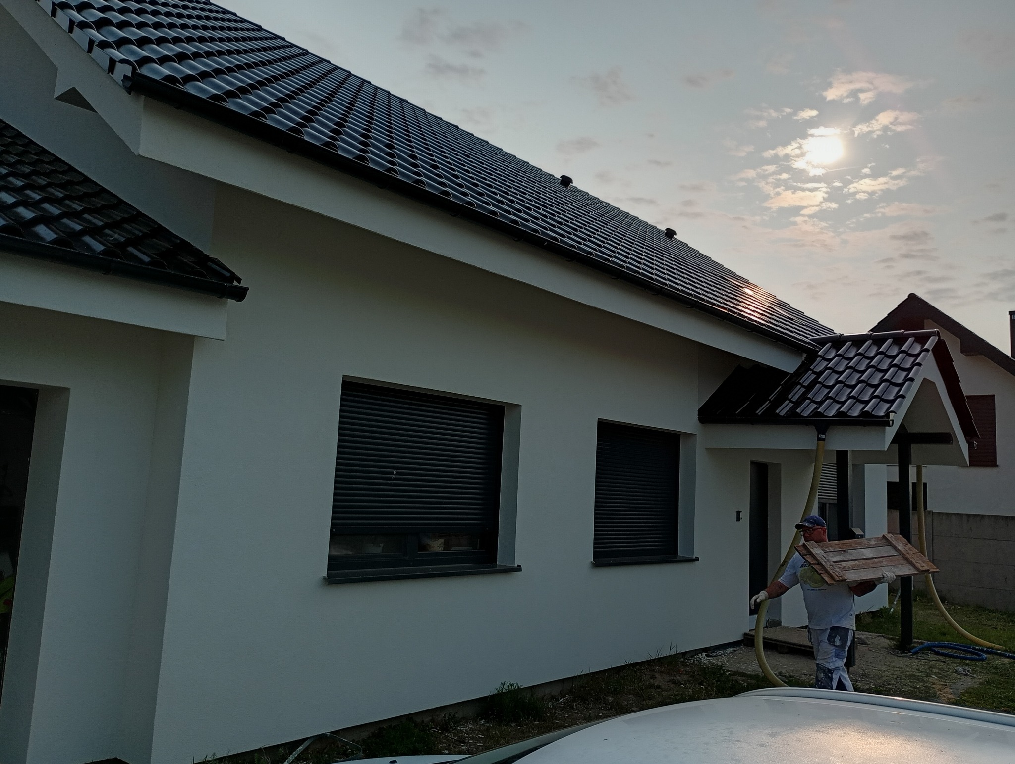 Biała elewacja domu jednorodzinnego z czarną dachówką, pracownik z deskami i wężem, słoneczny dzień. Widoczne okna z roletami zewnętrznymi i fragment dachu sąsiedniego budynku.