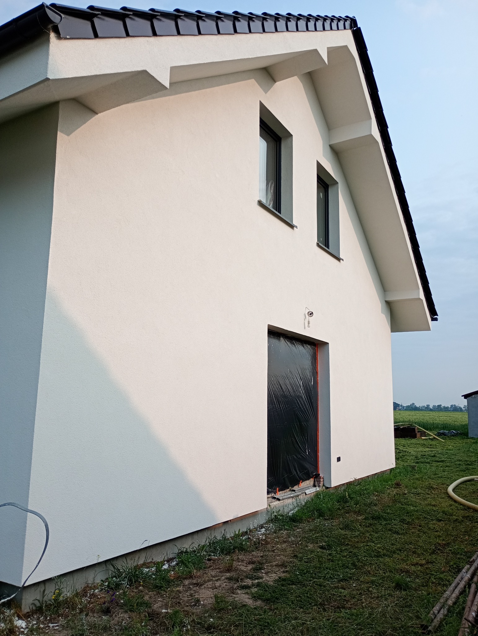 Boczny widok domu z białą elewacją, czarnym dachem i oknami. Wejście zabezpieczone folią. Widoczne trawnik i pola w tle. Ujęcie z dołu podkreśla geometrię budynku.