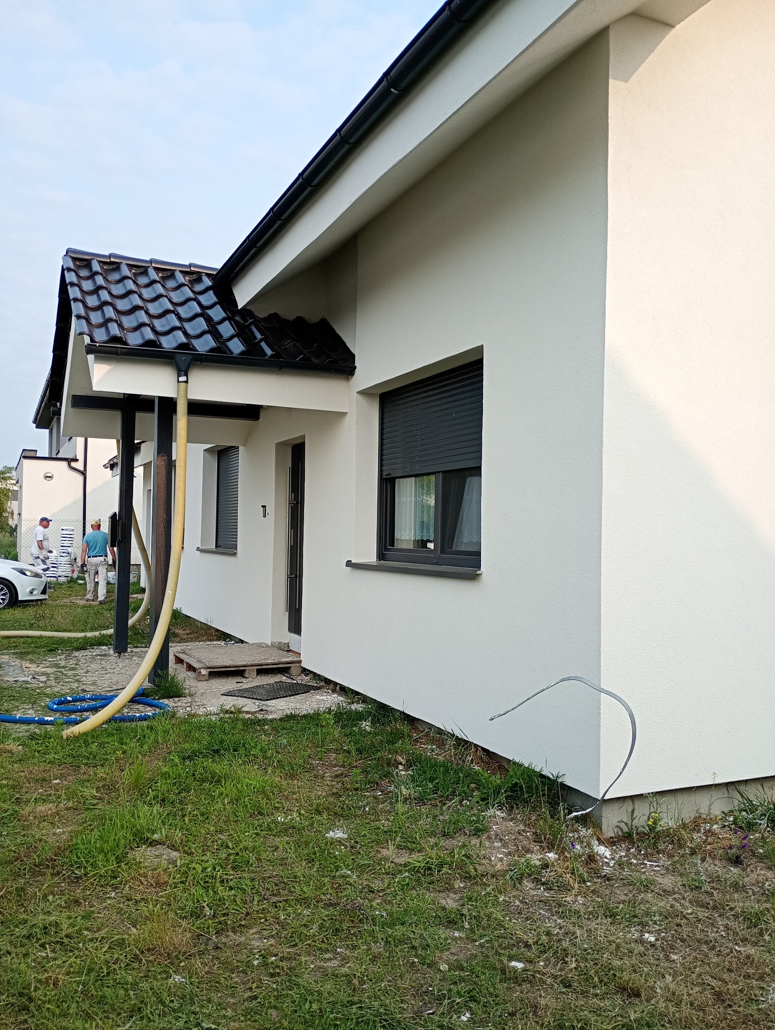 Biała elewacja domu jednorodzinnego z czarną dachówką, widoczni pracownicy w tle, żółty wąż do agregatu tynkarskiego. Świeżo położony tynk.