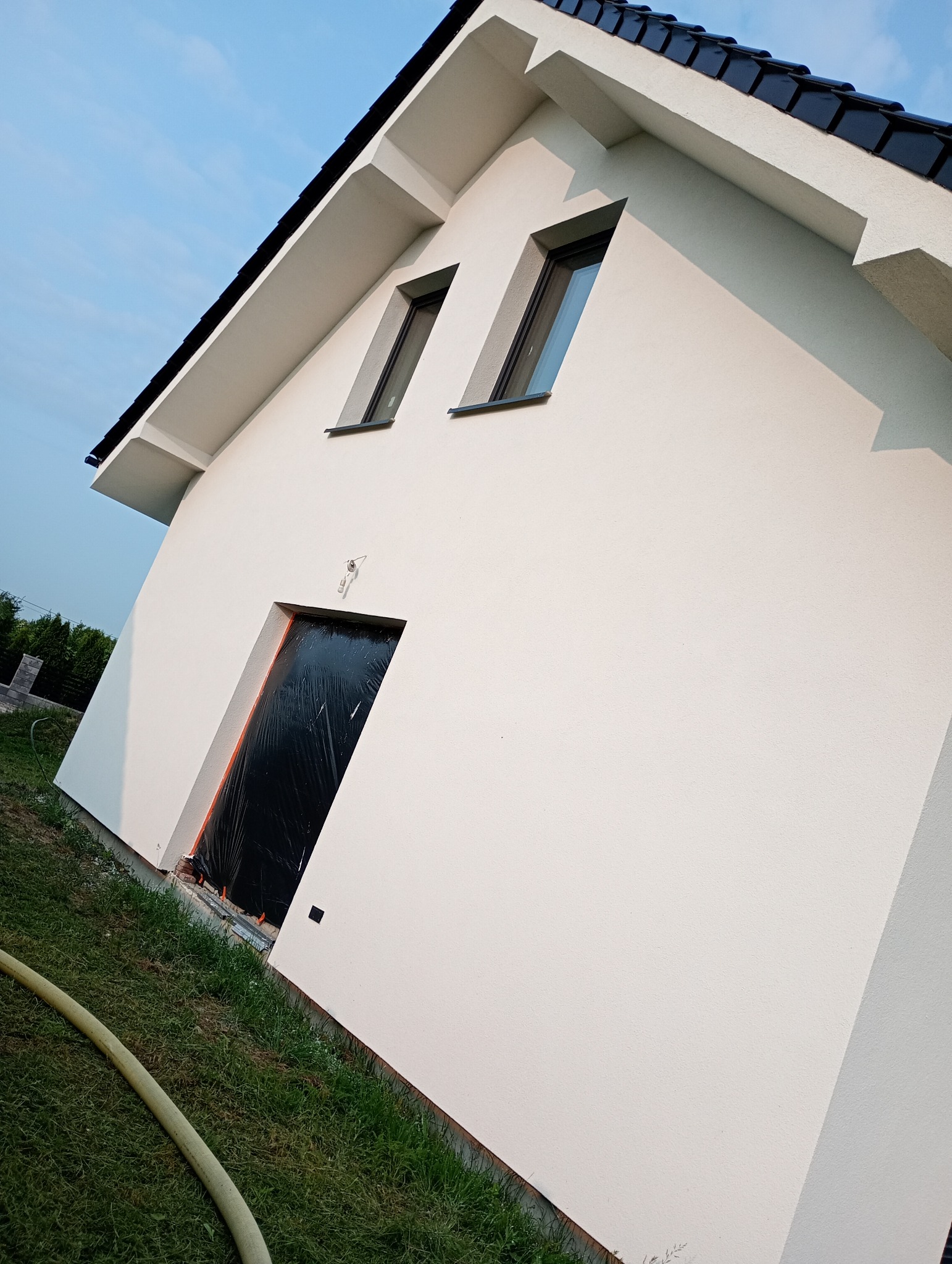 Biała elewacja domu jednorodzinnego z czarnym dachem i oknami. Wejście zabezpieczone folią. Ujęcie z dołu, podkreślające geometrię budynku i świeżość wykończenia.