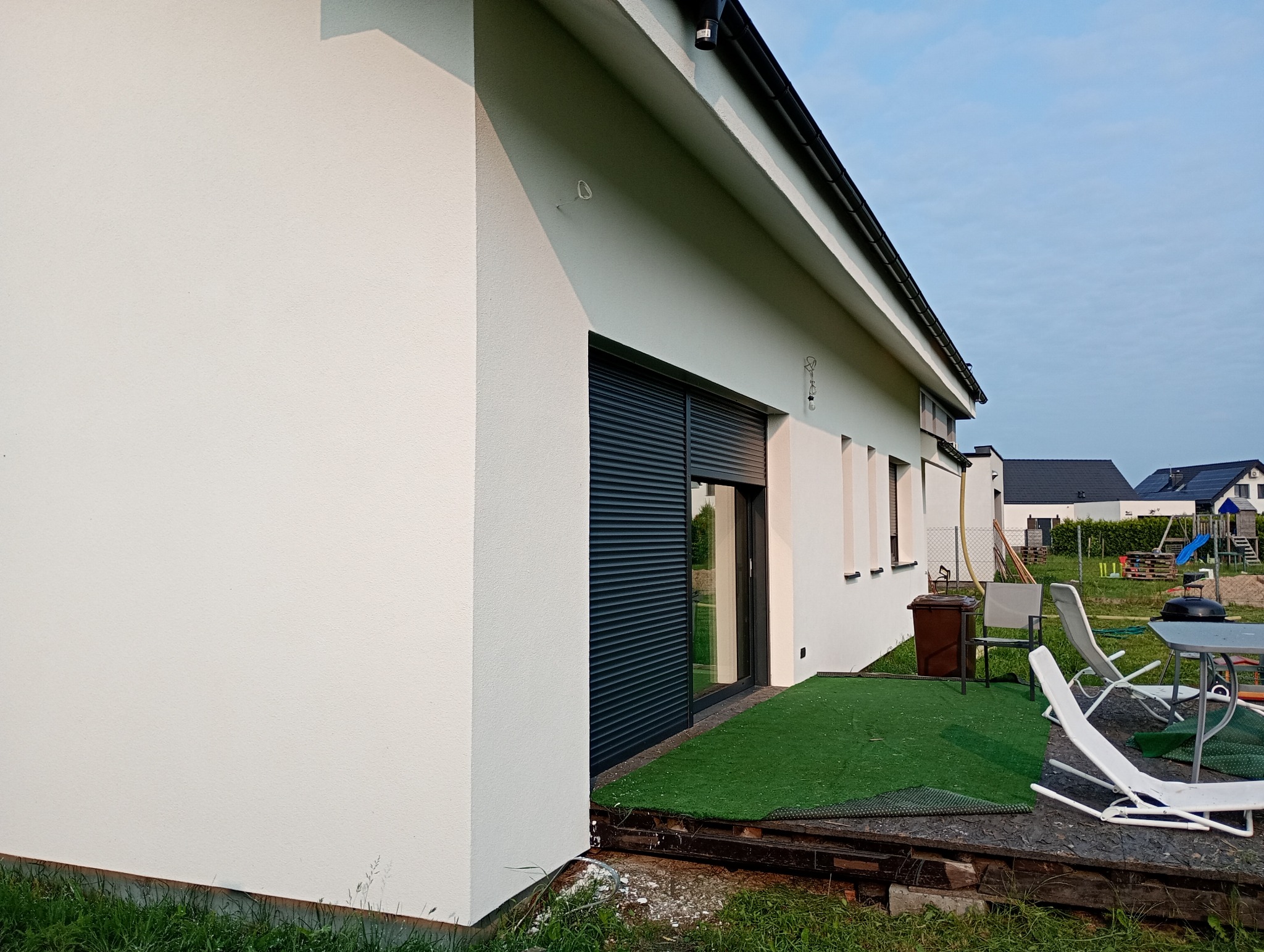 Biała elewacja domu jednorodzinnego z czarnymi roletami okiennymi i zielonym trawnikiem, widok z boku, w tle zabudowa sąsiedzka i plac zabaw.