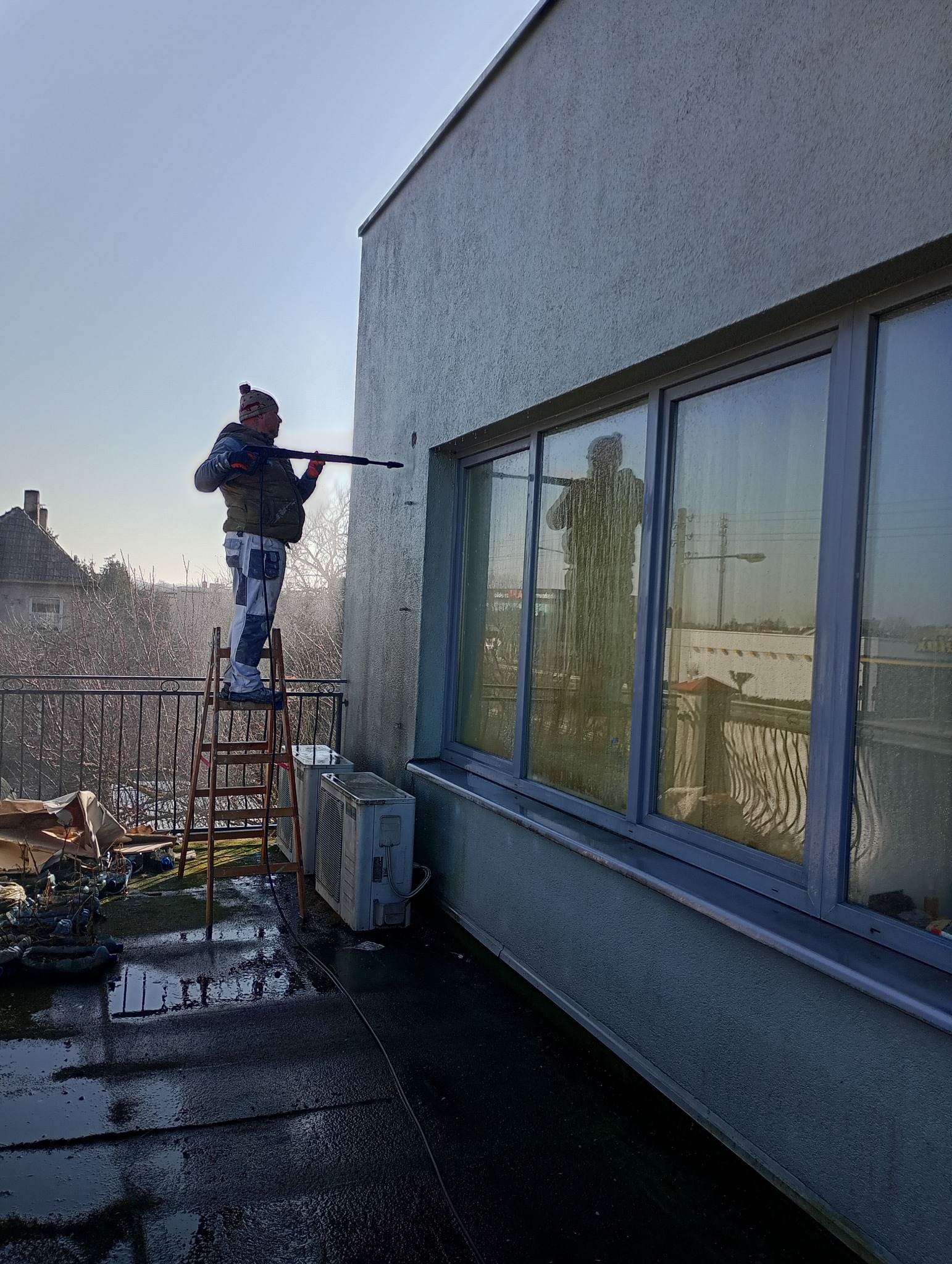 Pracownik na drabinie czyści elewację budynku myjką ciśnieniową. Widoczne okna z odbiciem sylwetki, mokry balkon i fragment sąsiedniego domu.
