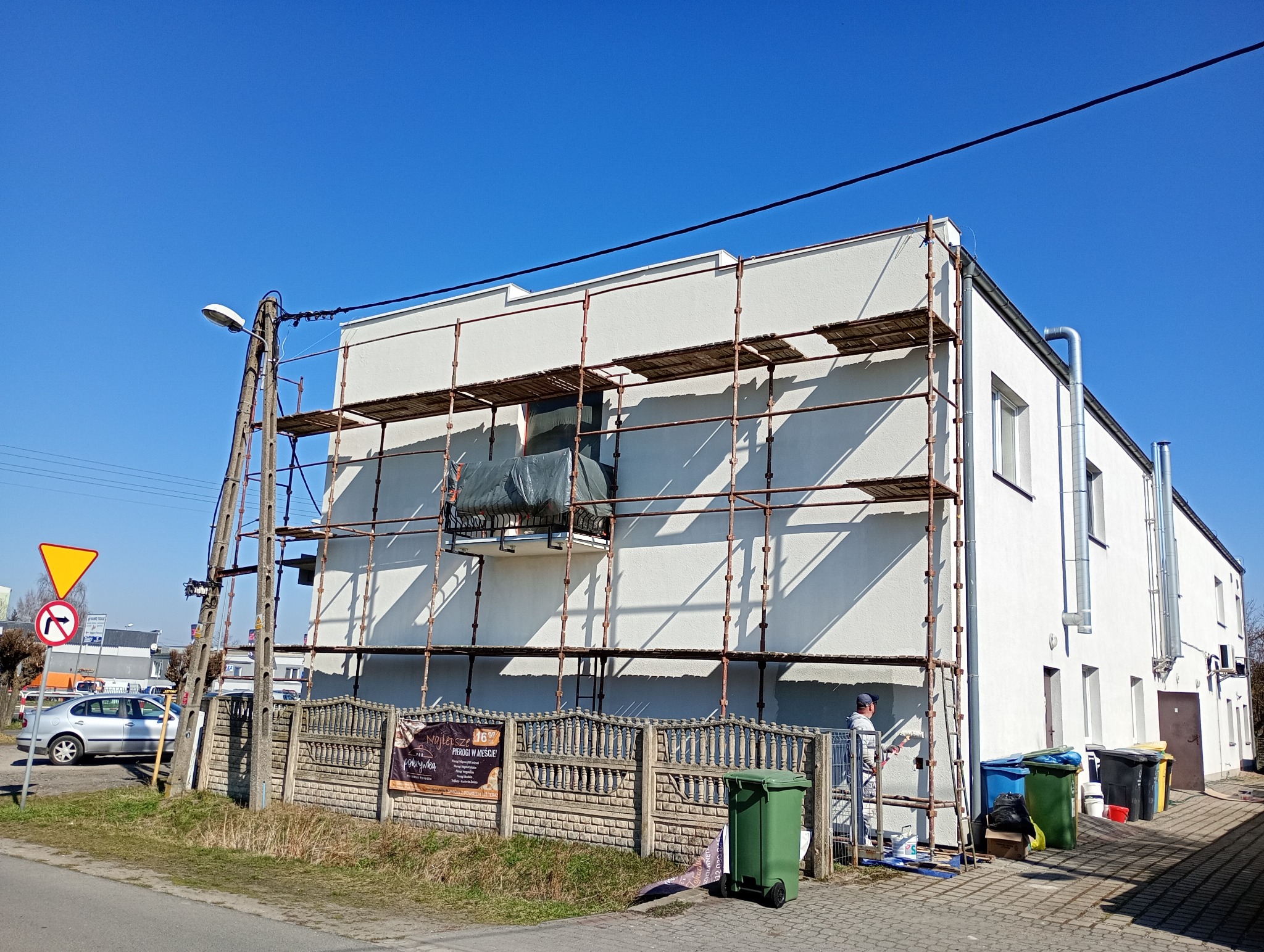 Biała elewacja budynku w trakcie malowania, widoczne rusztowania i pracownik z wałkiem. Słoneczny dzień, błękitne niebo. Prace wykończeniowe na zewnątrz.