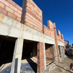 Wznoszenie ścian z pustaków ceramicznych na konstrukcji żelbetowej, widoczne zbrojenia i niebieska plandeka zabezpieczająca podłoże wewnątrz budynku.