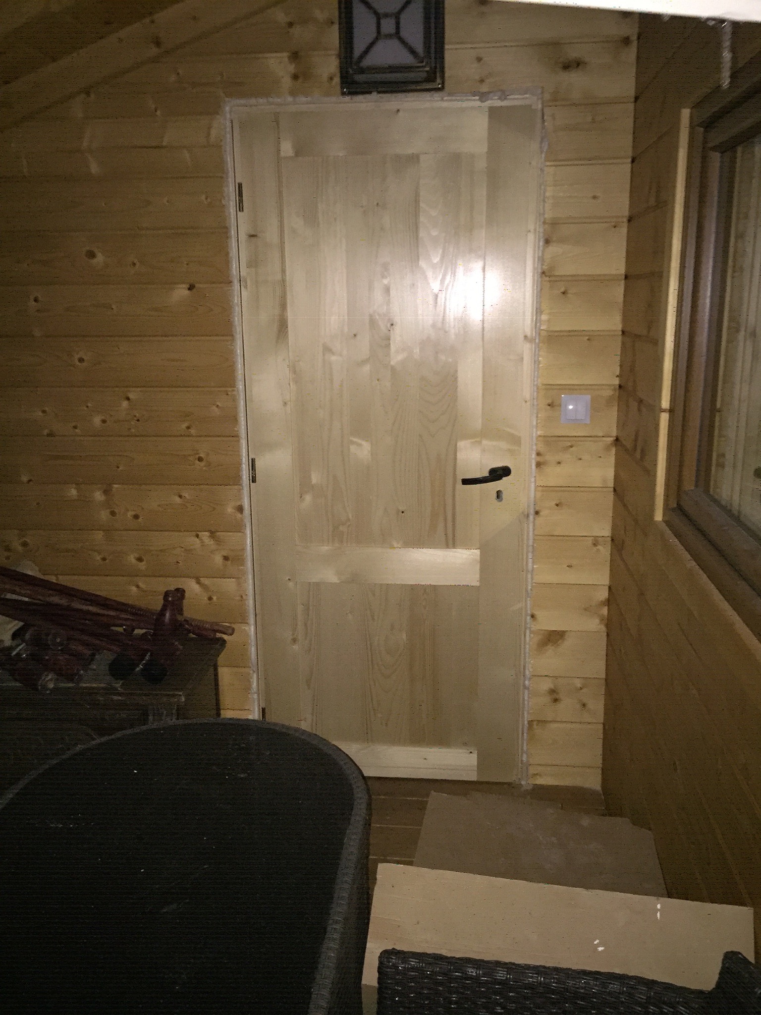 Surowa, niedokończona drewniana futryna i drzwi wewnątrz domku z boazerią, widoczny fragment okna i włącznik światła, na podłodze płyty chodnikowe.