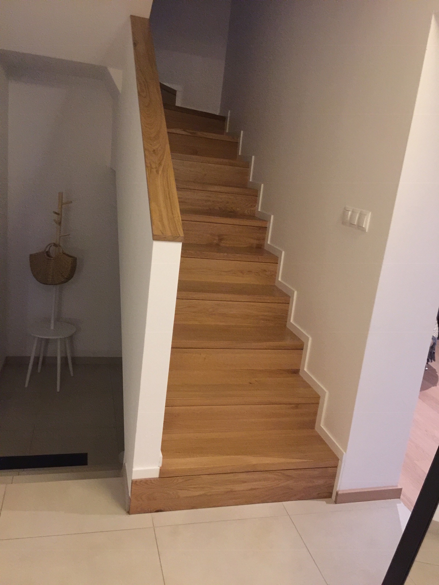 Nowoczesne, drewniane schody z jasnego drewna z białymi elementami wykończeniowymi przy ścianie, widok z dołu, w tle wieszak na ubrania.