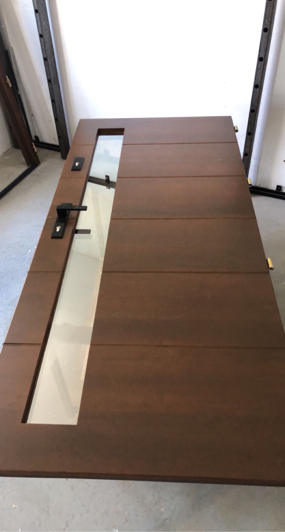 Ciemnobrązowe drzwi wewnętrzne z poziomymi panelami i prostokątną szybą, widoczne na podłodze w trakcie montażu lub obróbki.