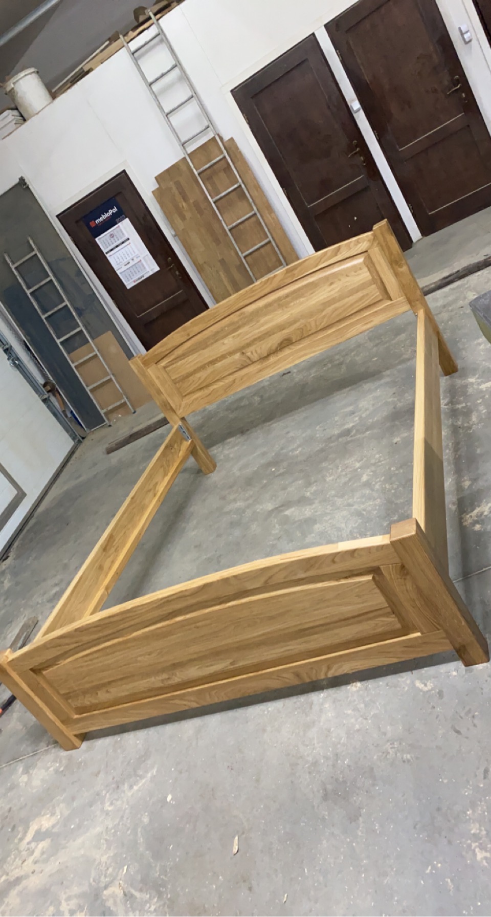 Surowa konstrukcja dębowa łóżka w warsztacie, widoczne drzwi w tle i drabina oparta o ścianę.