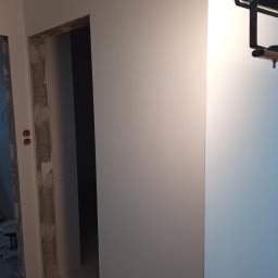 Usługi remontowe Marcin Ochałek - Świeżo pomalowana ściana w jasnym odcieniu szarości, widoczne surowe obramowanie drzwi przed wykończeniem, z instalacją elektryczną i lampą na ścianie.