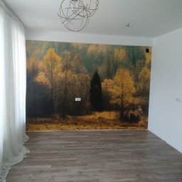 Usługi remontowe Marcin Ochałek - Pokój po remoncie z fototapetą przedstawiającą jesienny las, drewnopodobną podłogą, białymi ścianami i delikatną, ażurową firaną oraz nowoczesną lampą sufitową.