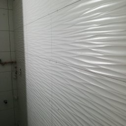 Usługi remontowe Marcin Ochałek - Białe, faliste płytki ścienne w trakcie montażu, widoczne krzyżyki dystansowe i poziomica, fragment instalacji wodnej po lewej stronie.