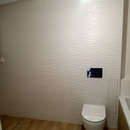 Usługi remontowe Marcin Ochałek - Nowoczesna toaleta podwieszana z przyciskiem spłukującym na ścianie wyłożonej trójwymiarowymi płytkami w odcieniach beżu, drewnopodobna podłoga.