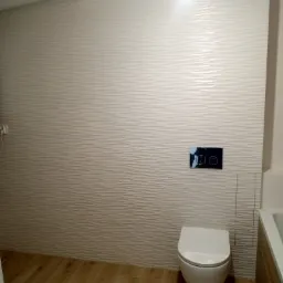 Nowoczesna toaleta podwieszana z przyciskiem spłukującym na ścianie wyłożonej trójwymiarowymi płytkami w odcieniach beżu, drewnopodobna podłoga.