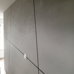 Usługi remontowe Marcin Ochałek - Ściana z szarą, teksturowaną powierzchnią, podzielona czarnymi, ukośnymi liniami tworzącymi geometryczny wzór. Widoczny biały włącznik światła i fragment podłogi z listwą przypodłogową.