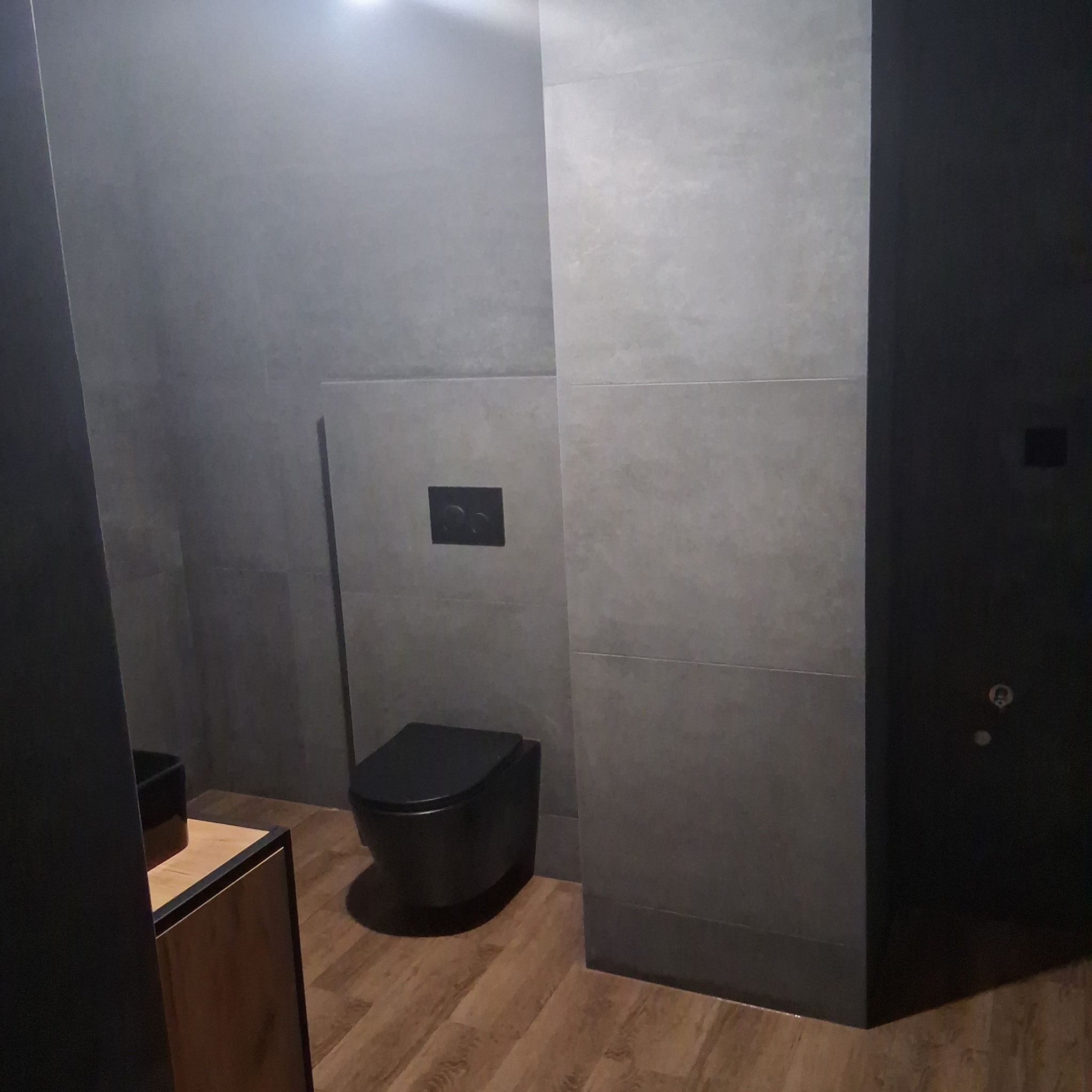 Nowoczesna łazienka w odcieniach szarości z czarną toaletą wiszącą i drewnopodobną podłogą, widoczny fragment szafki pod umywalkę z drewnianym frontem i czarnym blatem.