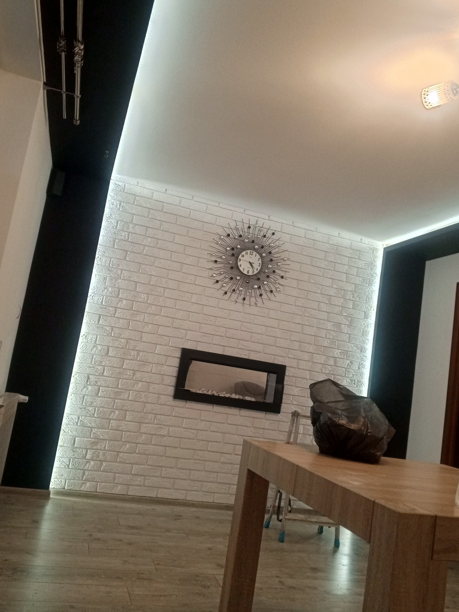 Wykończone wnętrze z białą cegłą na ścianie, ozdobnym zegarem, czarnym kominkiem i drewnianym stołem z zabezpieczonym przedmiotem, oświetlenie LED wzdłuż sufitu.