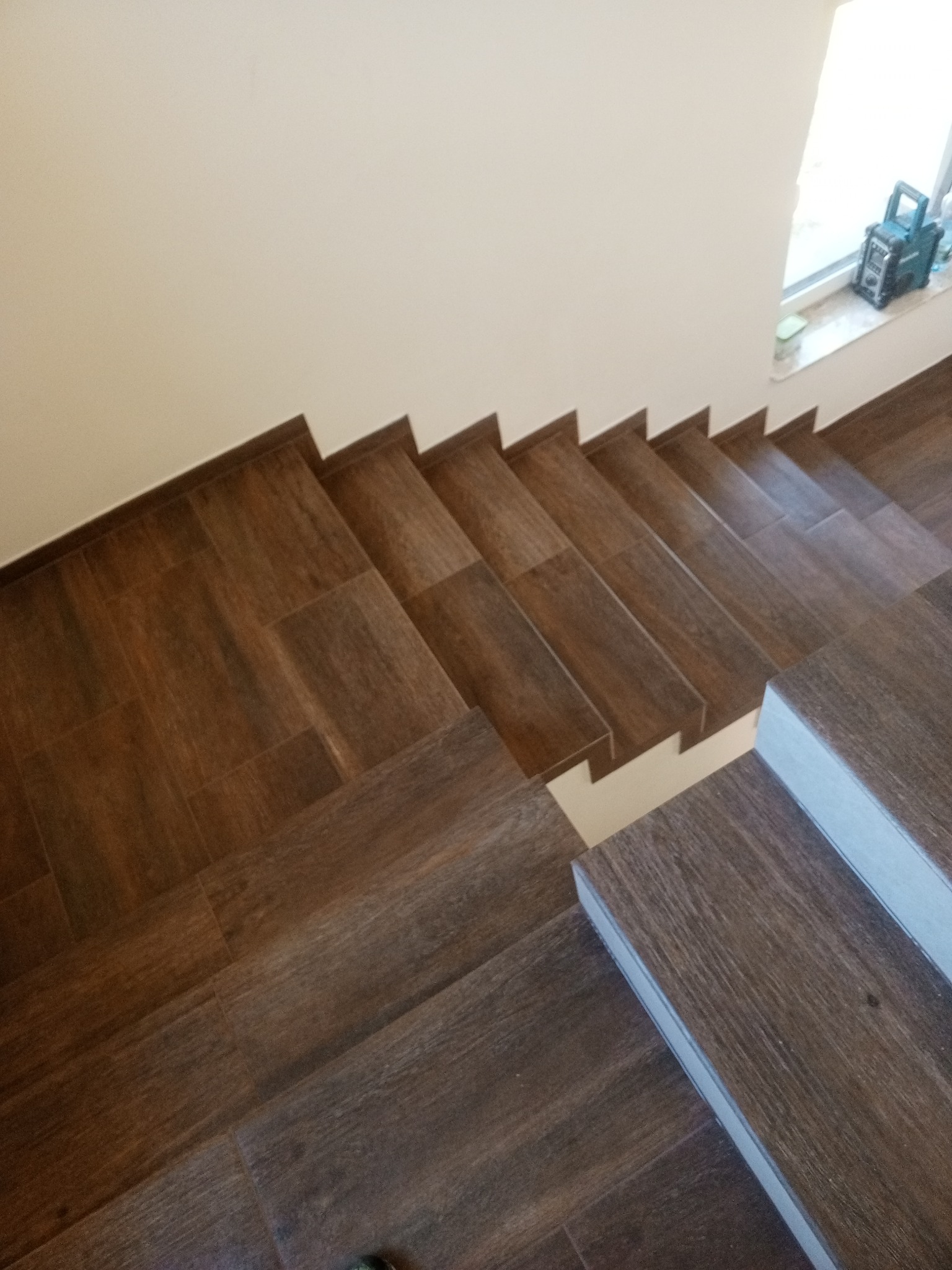 Ułożone płytki imitujące drewno na schodach wewnętrznych, widok z góry, z fragmentem ściany i parapetu z radiem budowlanym w tle.