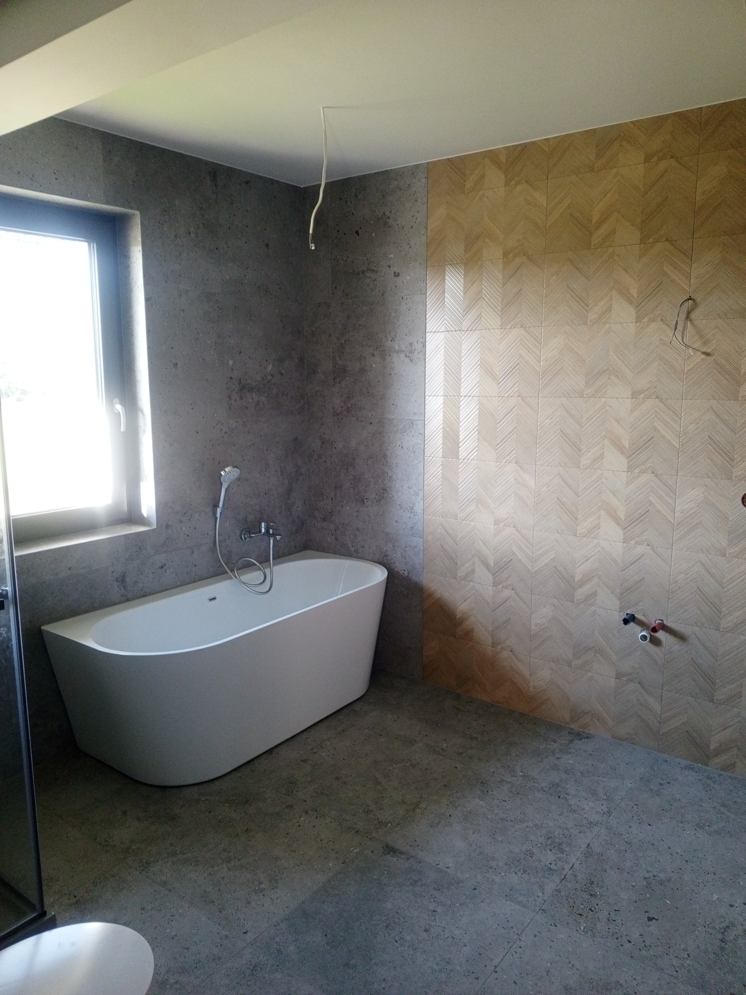Nowoczesna łazienka z wolnostojącą wanną, szarą betonową ścianą i podłogą oraz ścianą wyłożoną płytkami w jodełkę, widoczne wystające kable i rury.