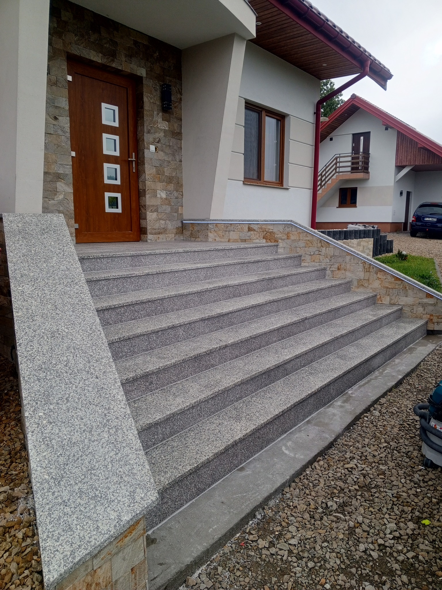 Granitowe schody wejściowe do domu z drewnianymi drzwiami i kamienną elewacją, widok z dołu na jasnoszare stopnie i rampę dla niepełnosprawnych.