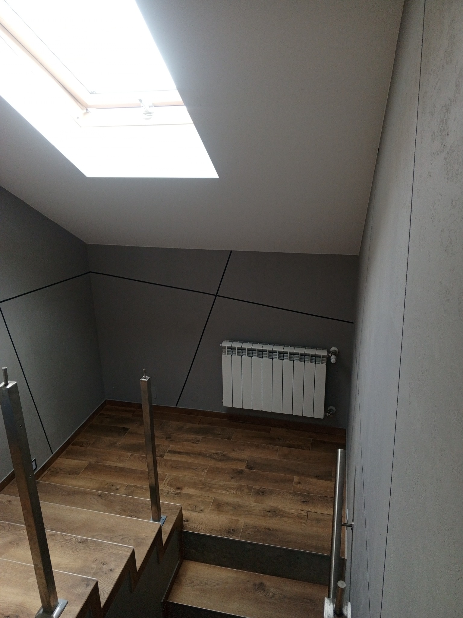 Wnętrze klatki schodowej z drewnopodobnymi płytkami na podłodze i schodach, szara ściana z dekoracyjnymi czarnymi liniami, biały grzejnik i okno dachowe w suficie.