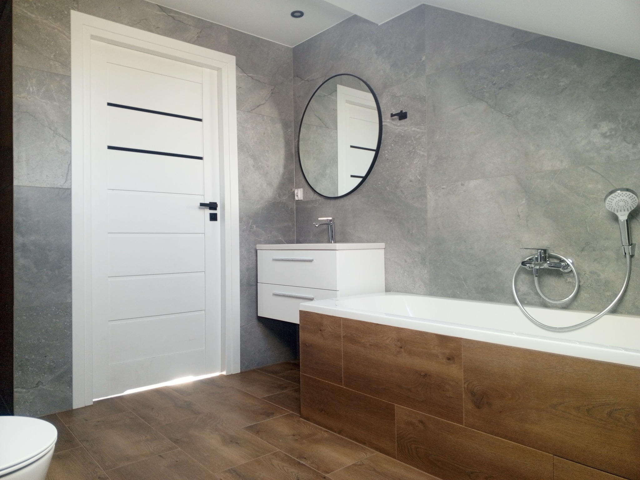 Nowoczesna łazienka z szarą marmurową płytką ścienną, drewnopodobną obudową wanny i podłogą, białymi meblami łazienkowymi i okrągłym lustrem.