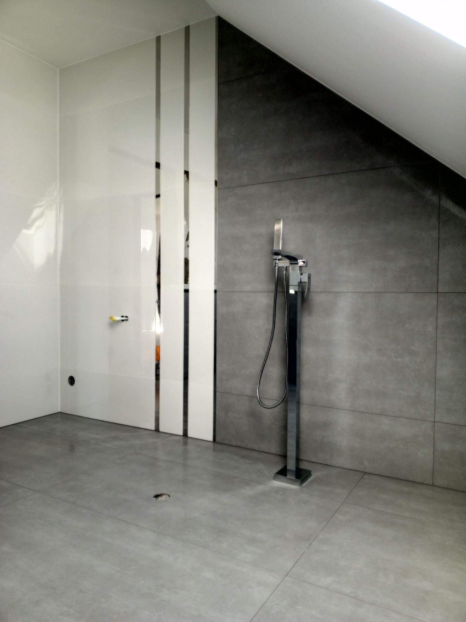 Nowoczesna łazienka na poddaszu z szarymi płytkami wielkoformatowymi na ścianie i podłodze, wolnostojąca chromowana bateria wannowa i biała ściana z lustrzanymi wstawkami.