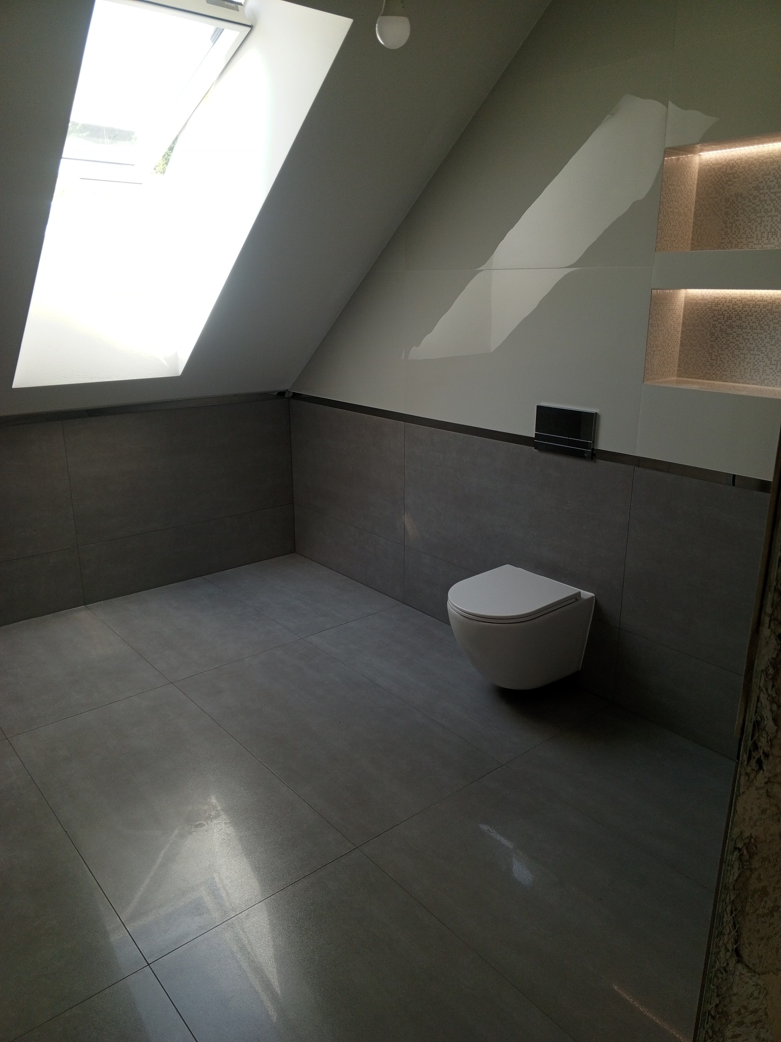 Nowoczesna łazienka na poddaszu z szarymi płytkami, podwieszaną toaletą i wnękami z mozaiką podświetloną LED, oświetlona światłem wpadającym przez okno dachowe.