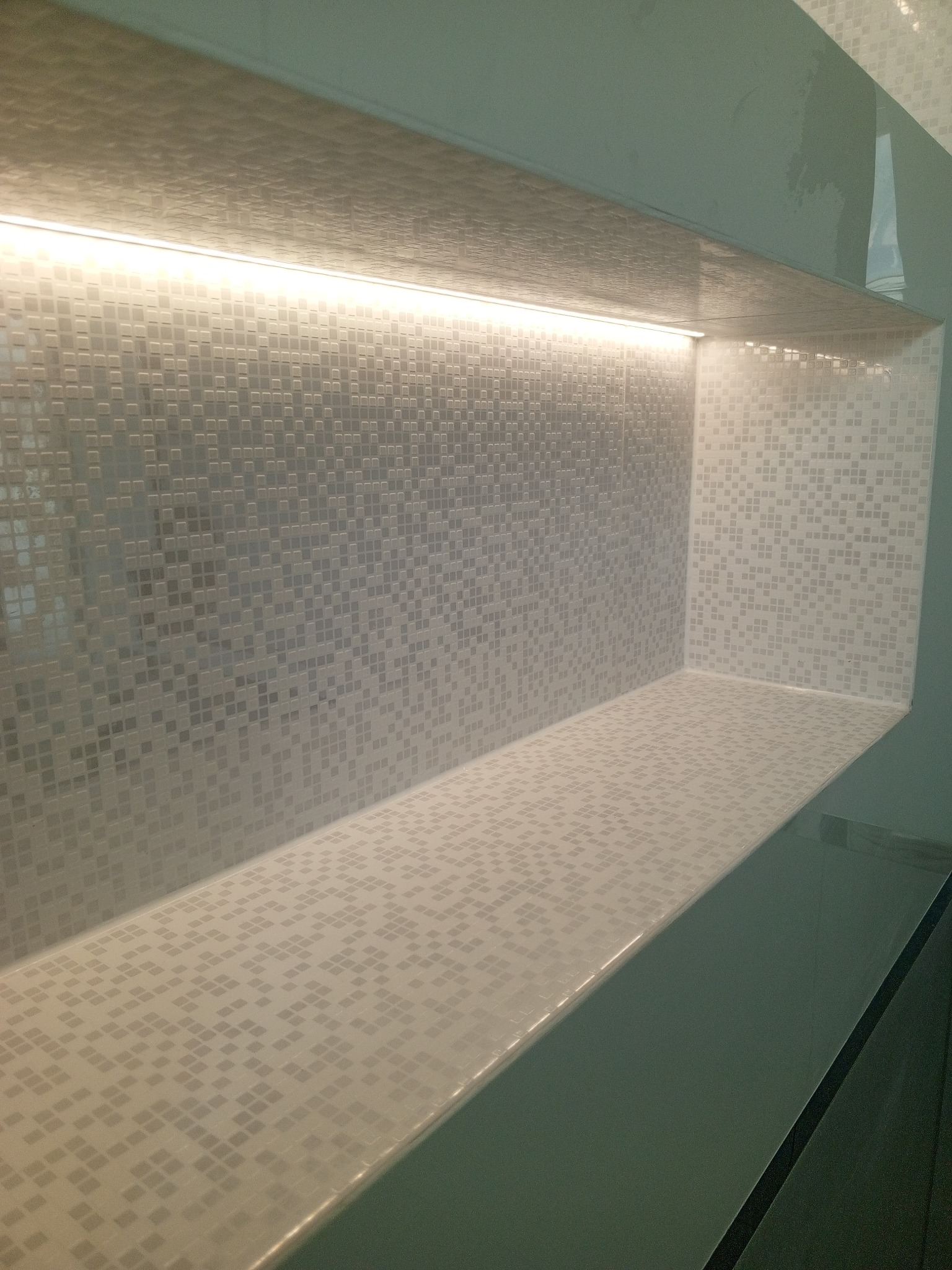 Półka wnękowa wyłożona mozaiką z drobnych, jasnych płytek, podświetlona listwą LED, widok z dołu.