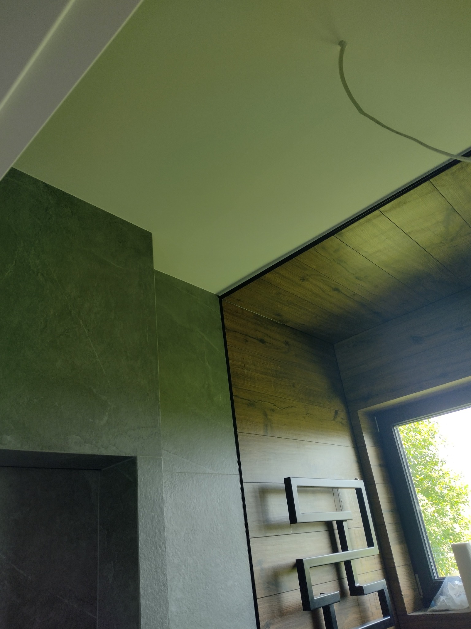 Fragment wnętrza łazienki z zielonymi płytkami imitującymi kamień na ścianie, drewnopodobną okładziną sufitu i ściany, oraz grzejnikiem drabinkowym w kolorze czarnym, widok z dołu na narożnik...