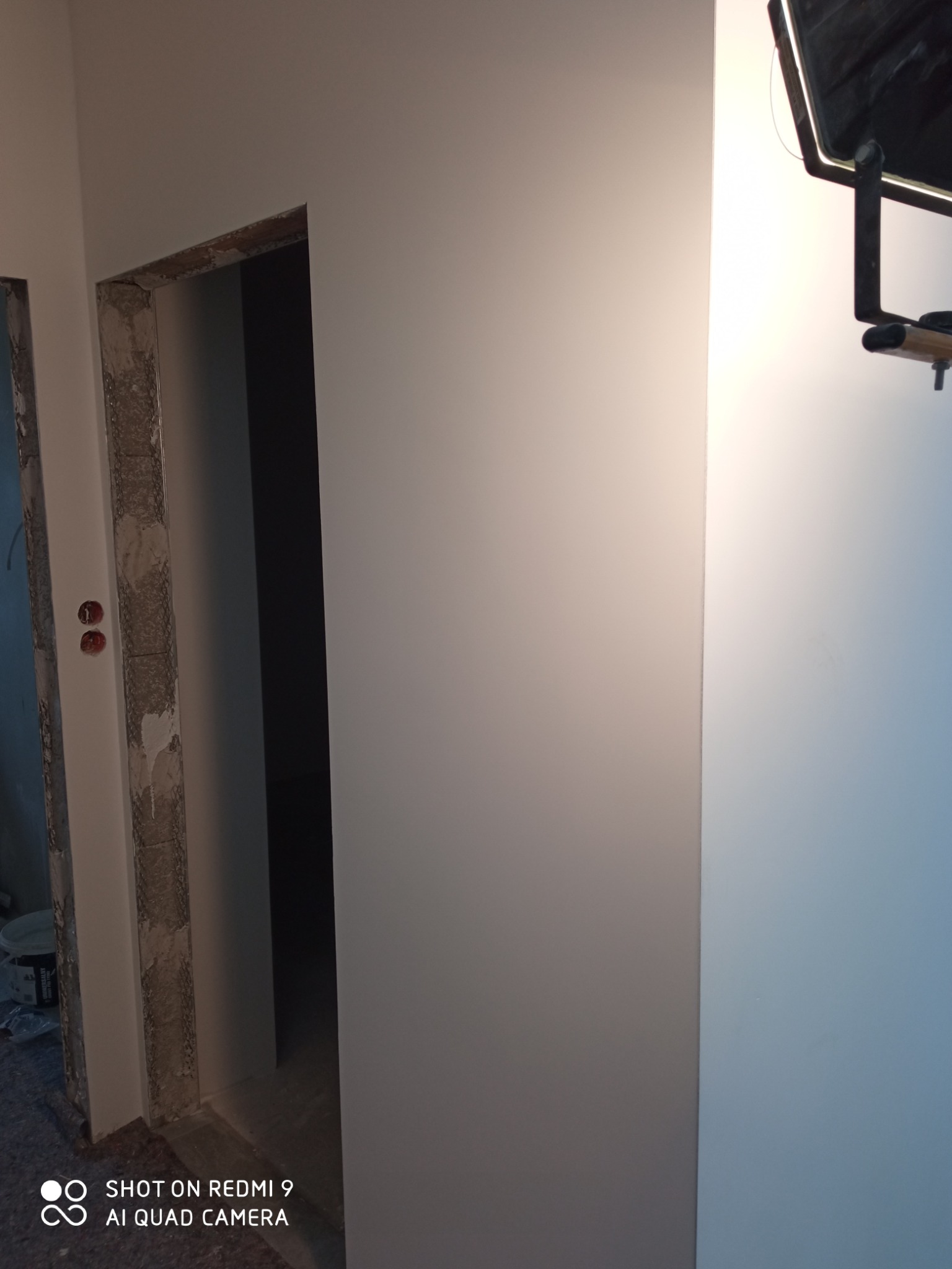 Świeżo pomalowana ściana w jasnym odcieniu szarości, widoczne surowe obramowanie drzwi przed wykończeniem, z instalacją elektryczną i lampą na ścianie.