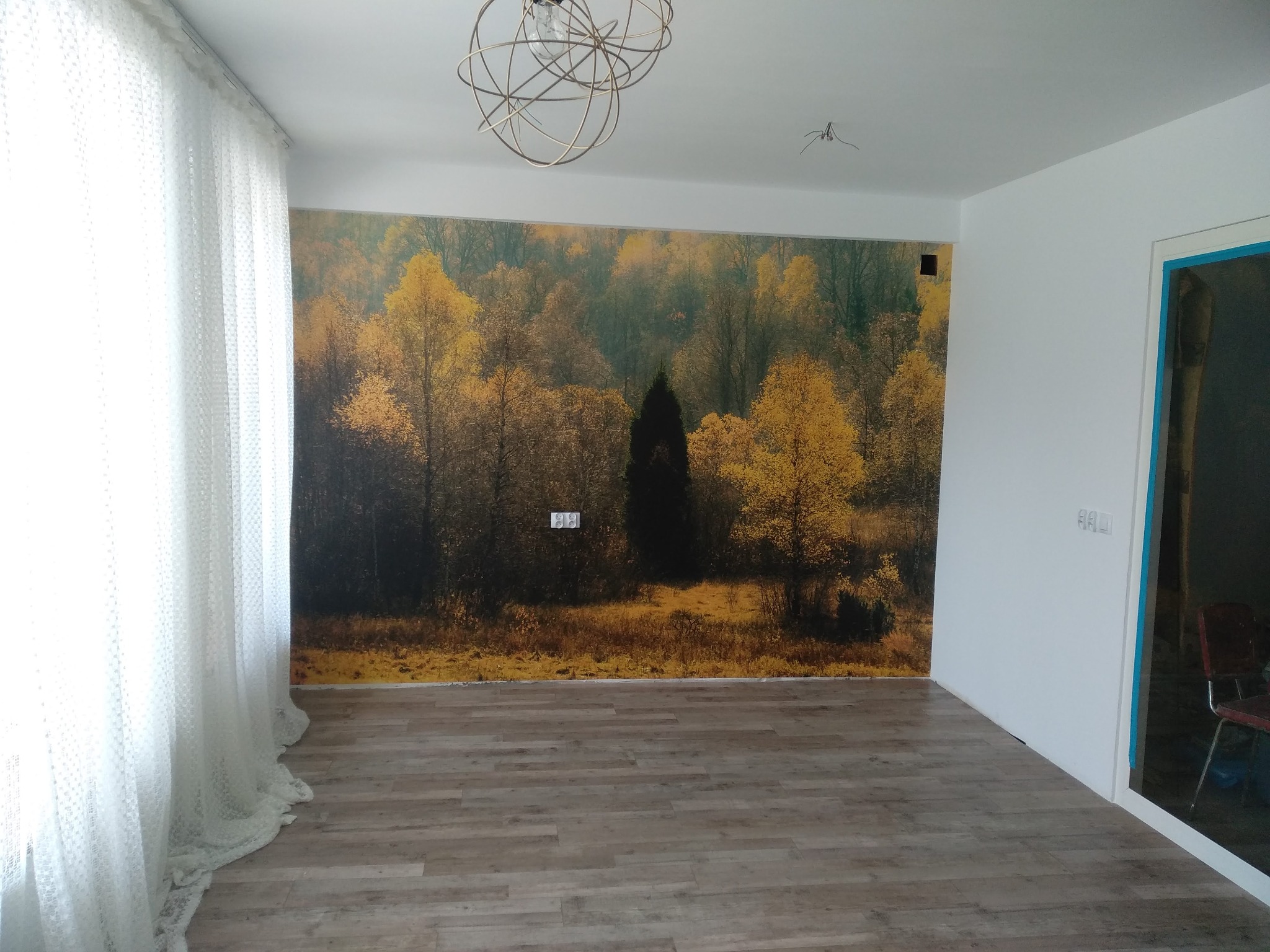 Pokój po remoncie z fototapetą przedstawiającą jesienny las, drewnopodobną podłogą, białymi ścianami i delikatną, ażurową firaną oraz nowoczesną lampą sufitową.