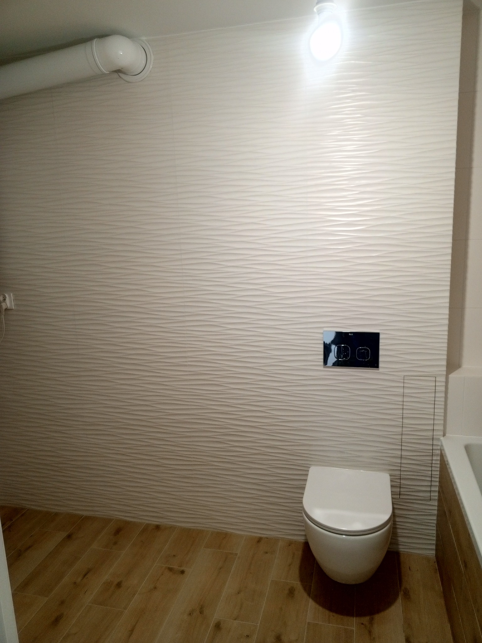 Nowoczesna toaleta podwieszana z przyciskiem spłukującym na ścianie wyłożonej trójwymiarowymi płytkami w odcieniach beżu, drewnopodobna podłoga.