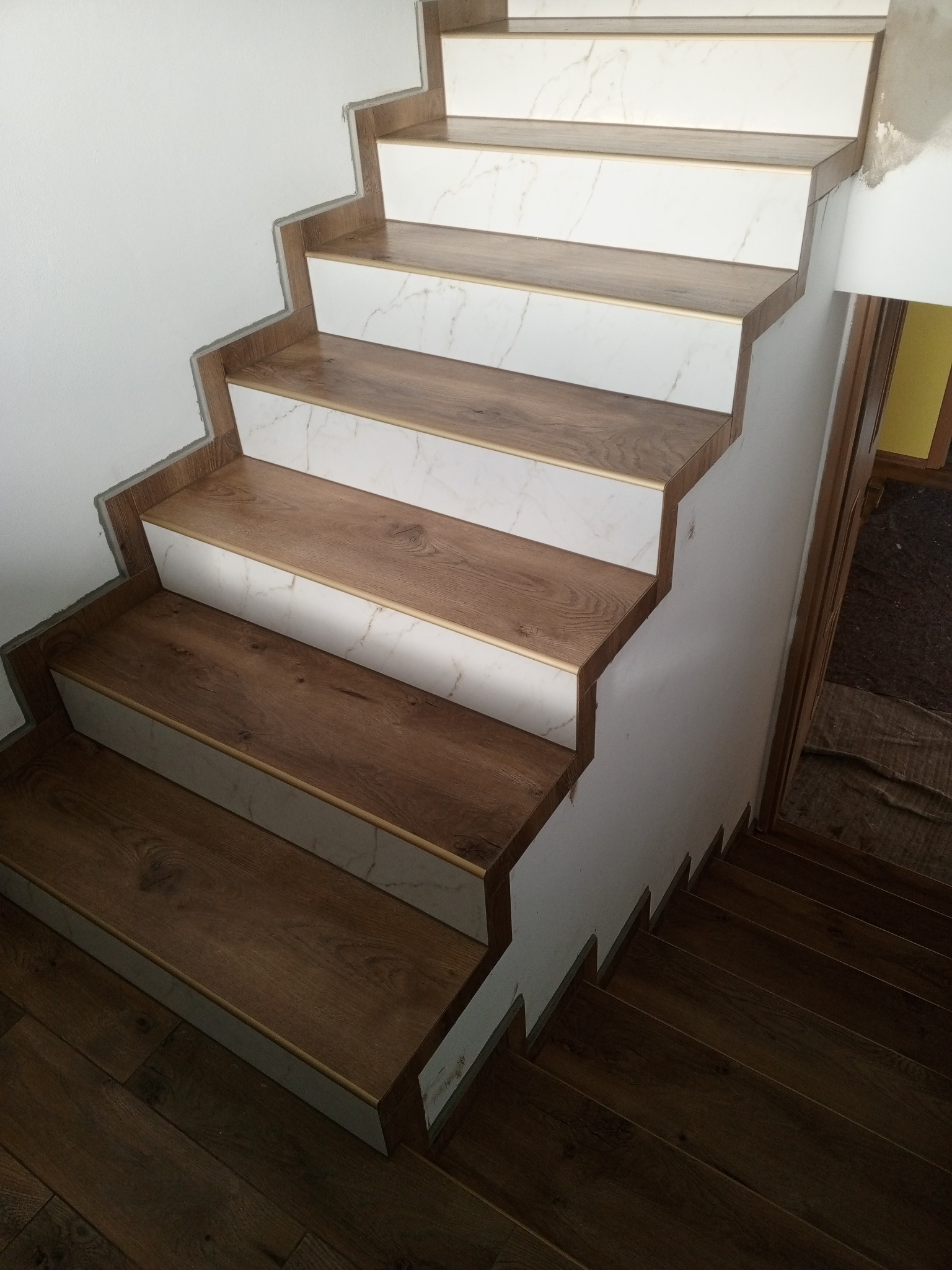 Schody wykończone panelami imitującymi drewno i białymi płytkami marmurowe, widoczne niedociągnięcia przy ścianie wymagające wykończenia.