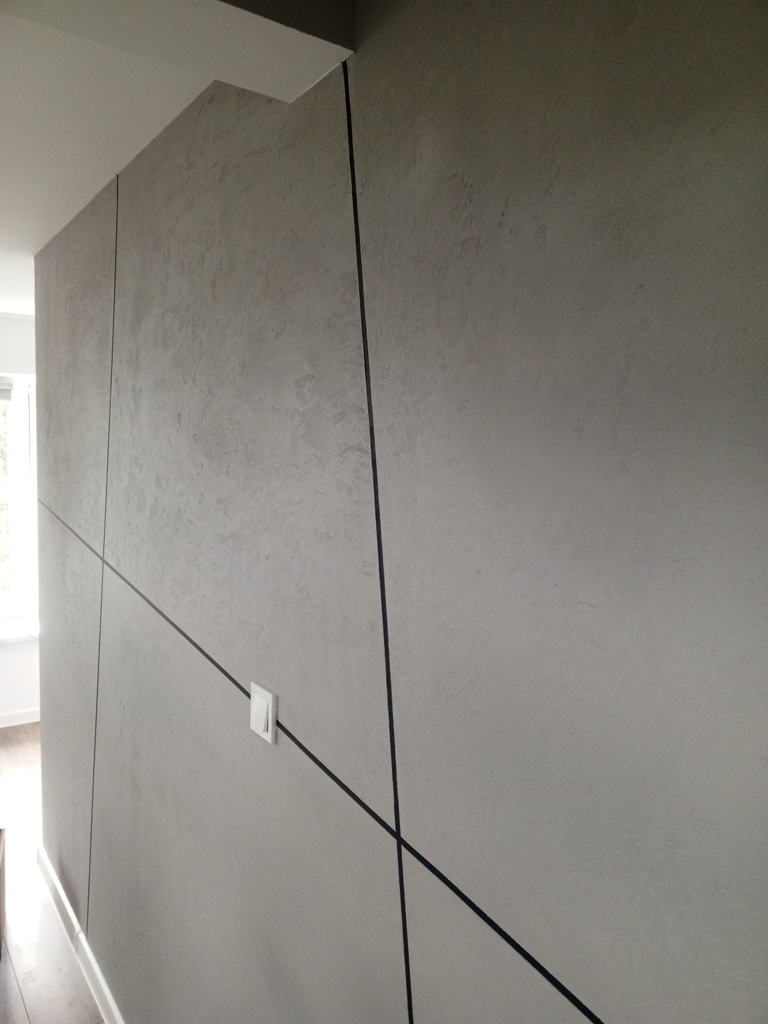 Ściana z szarą, teksturowaną powierzchnią, podzielona czarnymi, ukośnymi liniami tworzącymi geometryczny wzór. Widoczny biały włącznik światła i fragment podłogi z listwą przypodłogową.