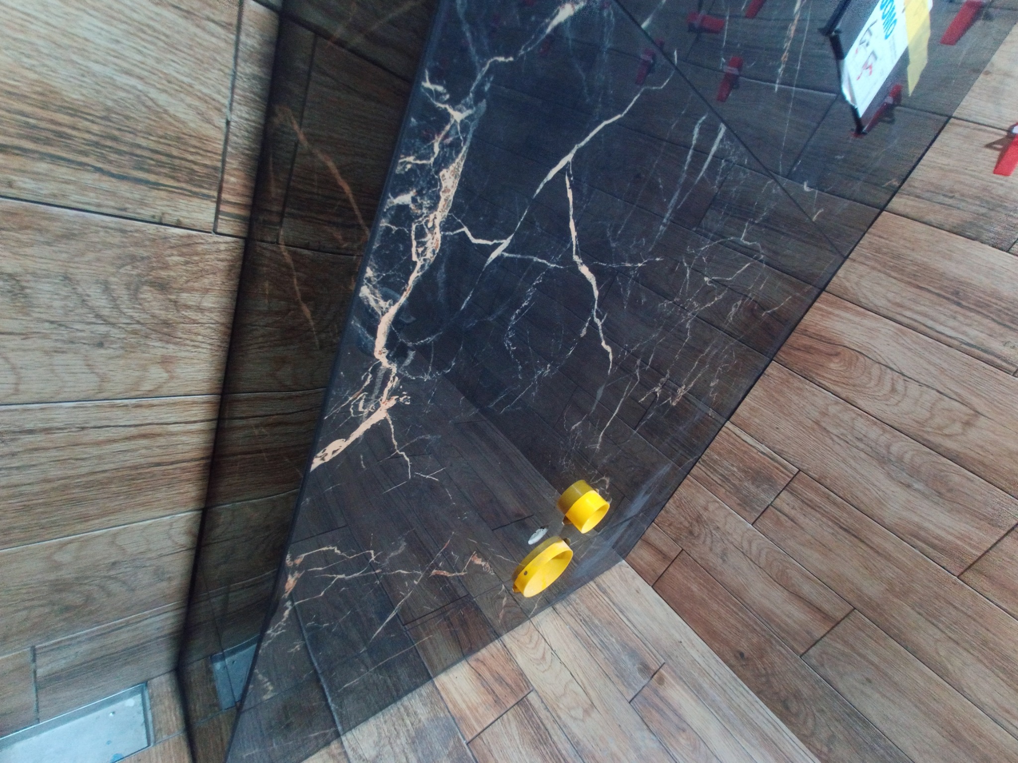Ułożona, błyszcząca, czarna płytka imitująca marmur z białymi żyłami, sąsiaduje z drewnopodobnymi płytkami. Widoczne żółte elementy poziomowania płytek i czerwone kliny.