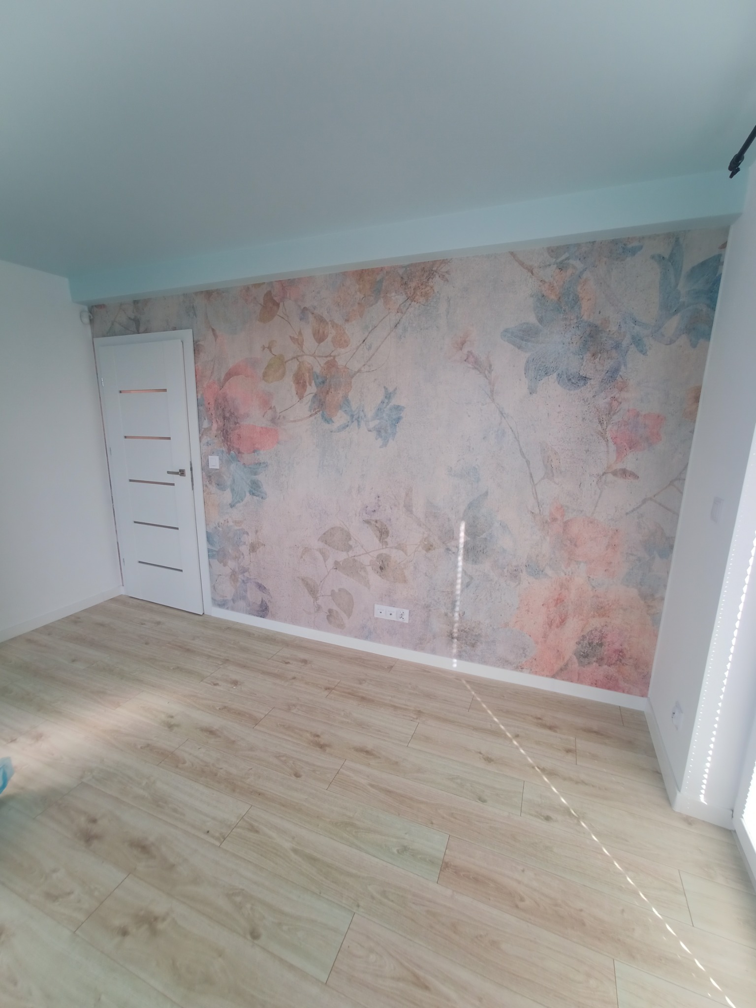 Pusty pokój po remoncie z nową tapetą w kwiaty, drewnianą podłogą i pomalowanym na jasny niebieski kolor sufitem.