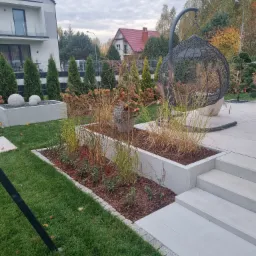 Nowoczesny ogród z podwyższonymi rabatami w betonowych obudowach, ozdobnymi kulami, trawami ozdobnymi i wiszącym fotelem kokonem na tle domu i jesiennego krajobrazu.