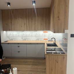 Home Koncept Rafal Magiera - Nowoczesna kuchnia z drewnianymi szafkami górnymi i szarymi dolnymi, marmurowy backsplash, czarny zlew i płyta indukcyjna, widoczna rolka papieru i skrzynka z narzędziami na podłodze.