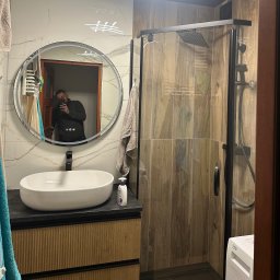 Home Koncept Rafal Magiera - Nowoczesna łazienka z okrągłym lustrem z podświetleniem LED, kabiną prysznicową typu walk-in z czarną armaturą, umywalką nablatową na szafce z drewnianymi lamelami i marmurową okładziną ścienną...