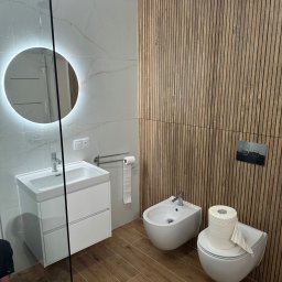Home Koncept Rafal Magiera - Nowoczesna łazienka z białymi płytkami imitującymi marmur, drewnianą ścianą z lameli, okrągłym lustrem z podświetleniem LED i podłogą z płytek imitujących drewno.