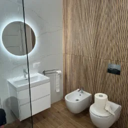 Nowoczesna łazienka z białymi płytkami imitującymi marmur, drewnianą ścianą z lameli, okrągłym lustrem z podświetleniem LED i podłogą z płytek imitujących drewno.