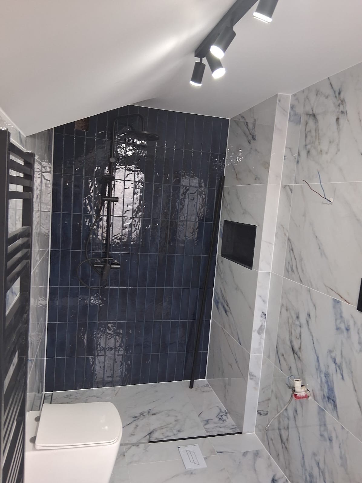 Nowoczesna łazienka z kabiną prysznicową wyłożoną pionowymi, ciemnoniebieskimi płytkami, marmurowe płytki na ścianach i podłodze, czarna armatura i oświetlenie punktowe, widoczny fragment toalety...