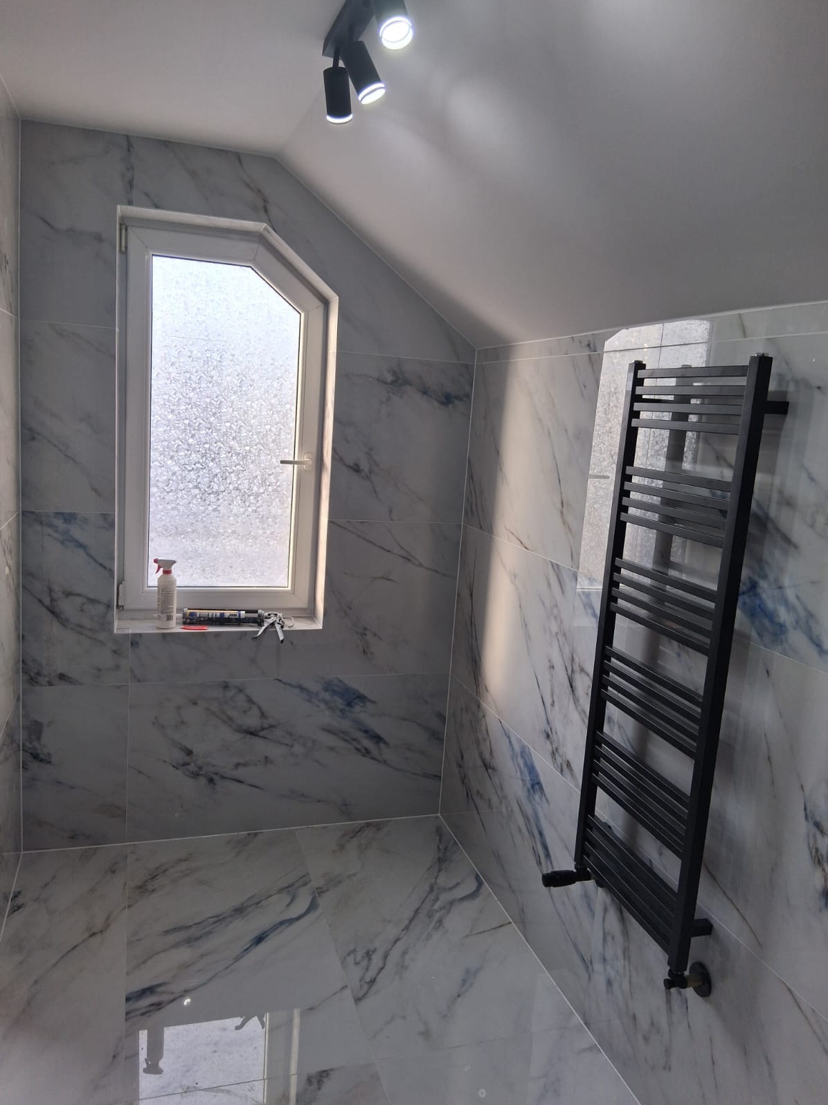 Wykończona łazienka na poddaszu z marmurowymi płytkami, czarnym grzejnikiem drabinkowym i oknem ze spryskiwaną szybą, na parapecie butelka i pistolet do silikonu.