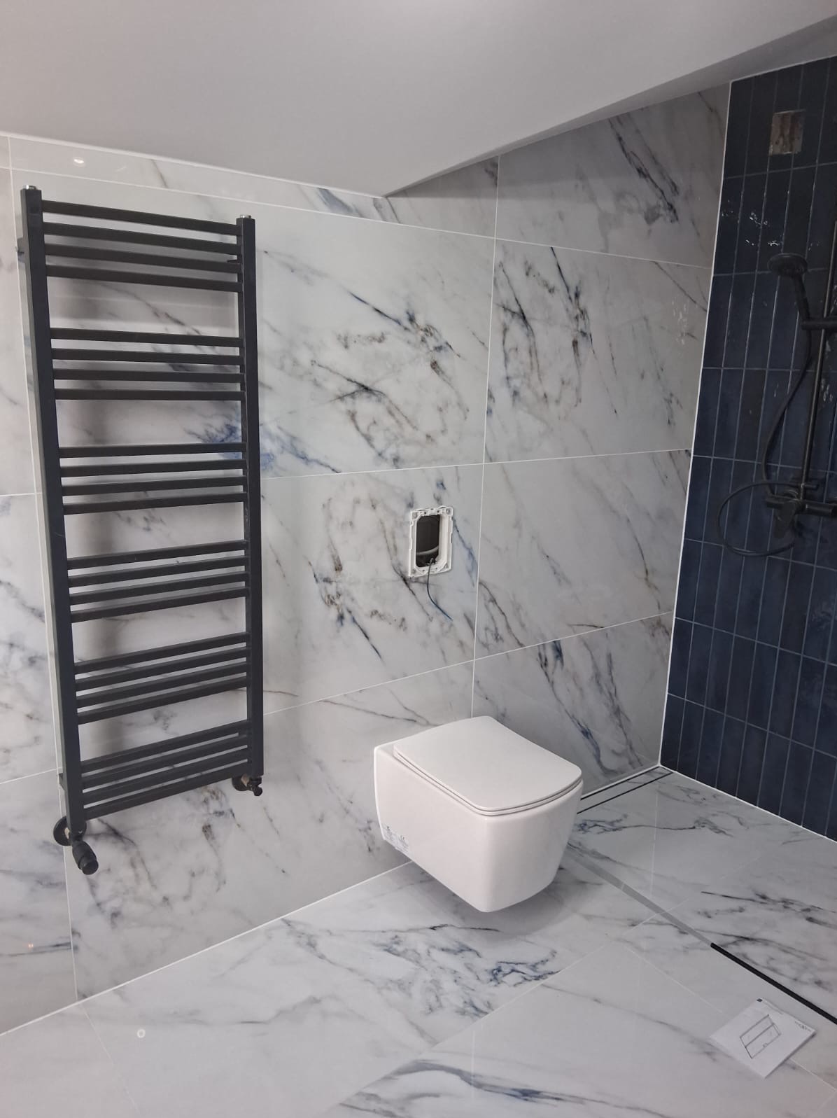 Nowoczesna łazienka z marmurowymi płytkami, wisząca toaleta i czarny grzejnik drabinkowy na kółkach.
