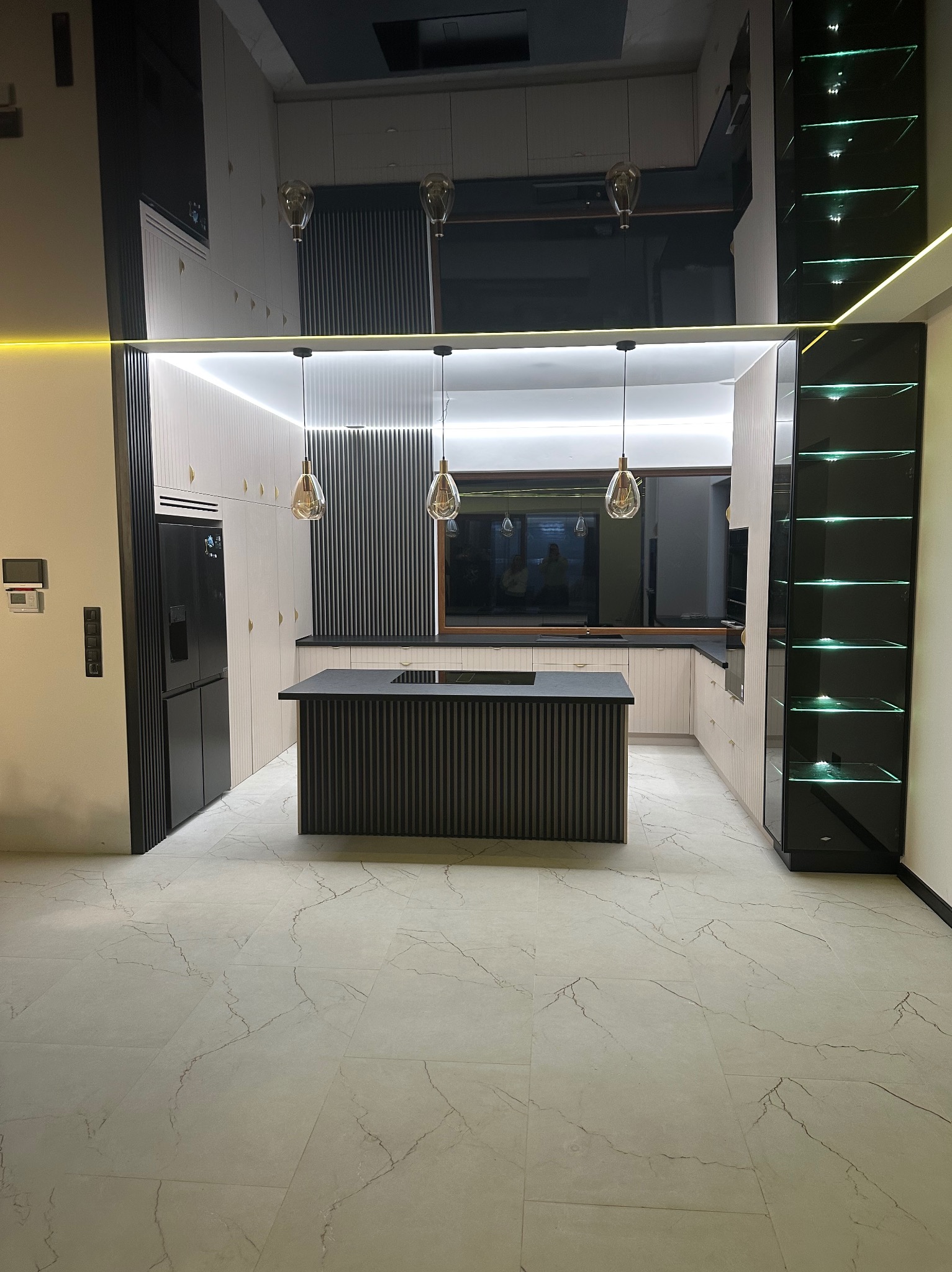 Nowoczesna kuchnia z wyspą na marmurowej podłodze, oświetlona wiszącymi lampami i podświetleniem LED, zabudowa w kolorach bieli i czerni z pionowymi listwami.