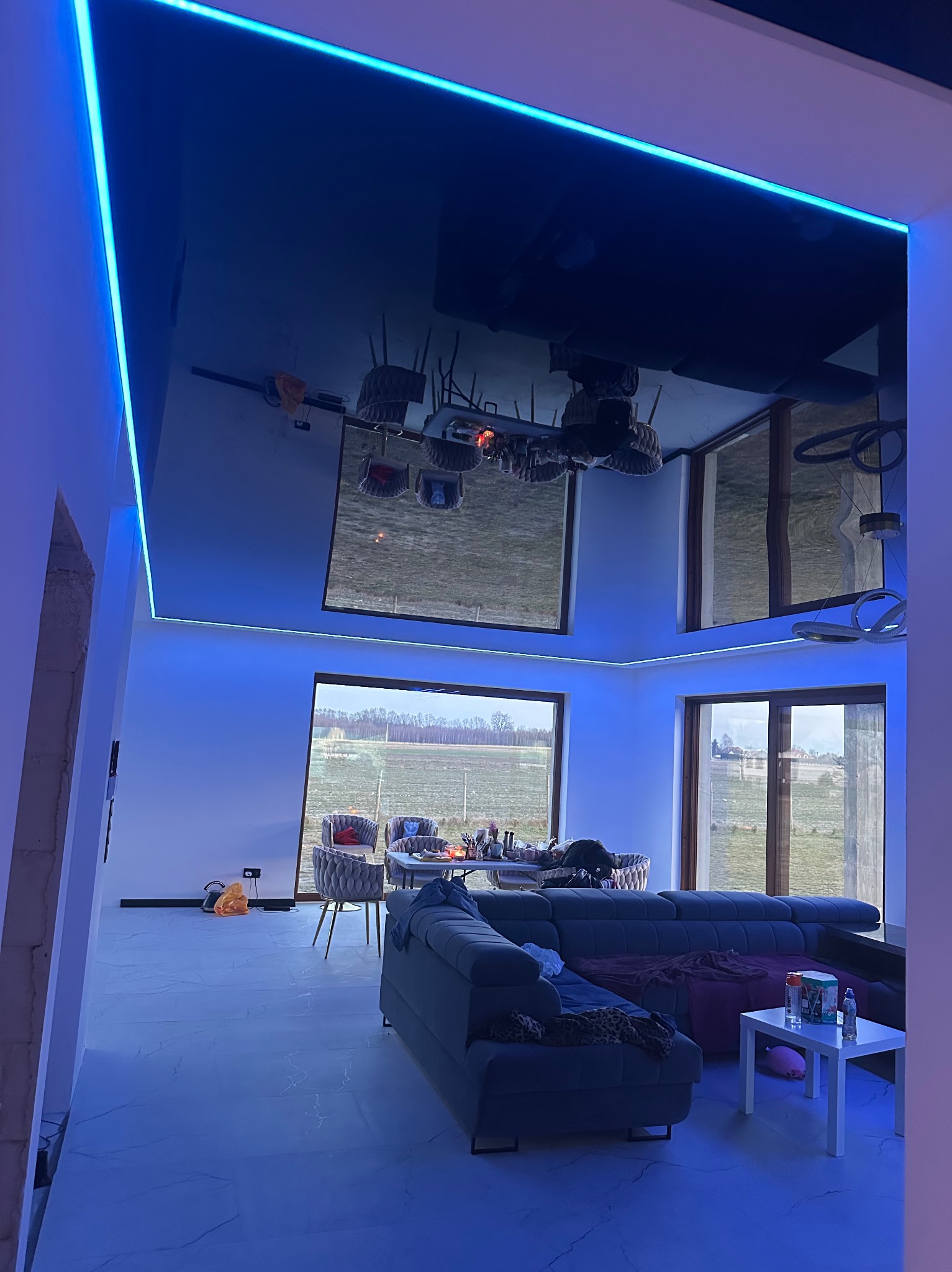 Nowoczesny salon z czarnym, błyszczącym sufitem podwieszanym, oświetlonym niebieskim LED, duże okna z widokiem na zewnątrz, szara sofa i stół z krzesłami w tle.