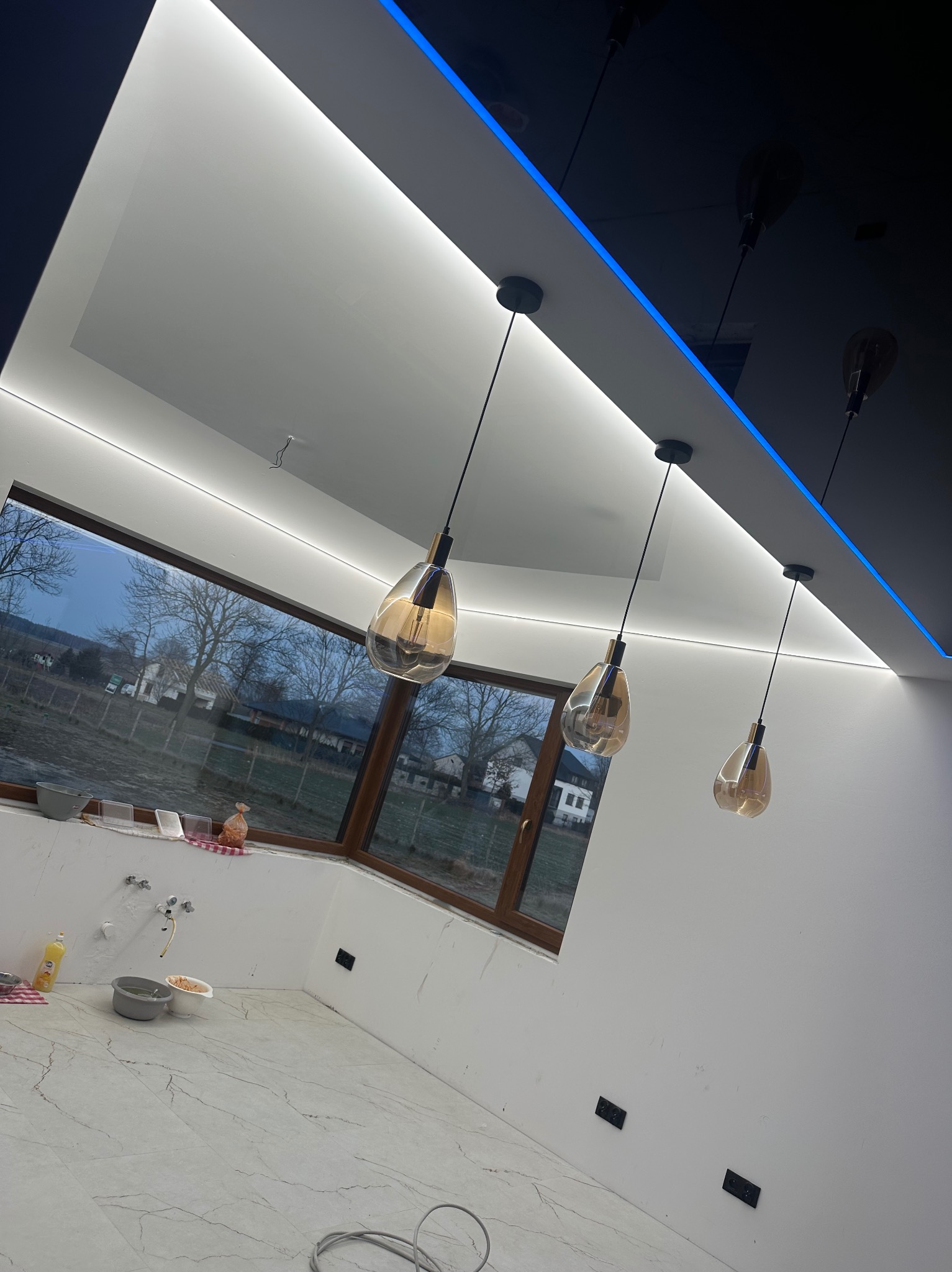 Wnętrze łazienki w trakcie remontu: białe ściany przygotowane do malowania, marmurowe płytki podłogowe, okno z widokiem na zewnątrz, trzy wiszące lampy z brązowymi kloszami, podwieszany sufit...