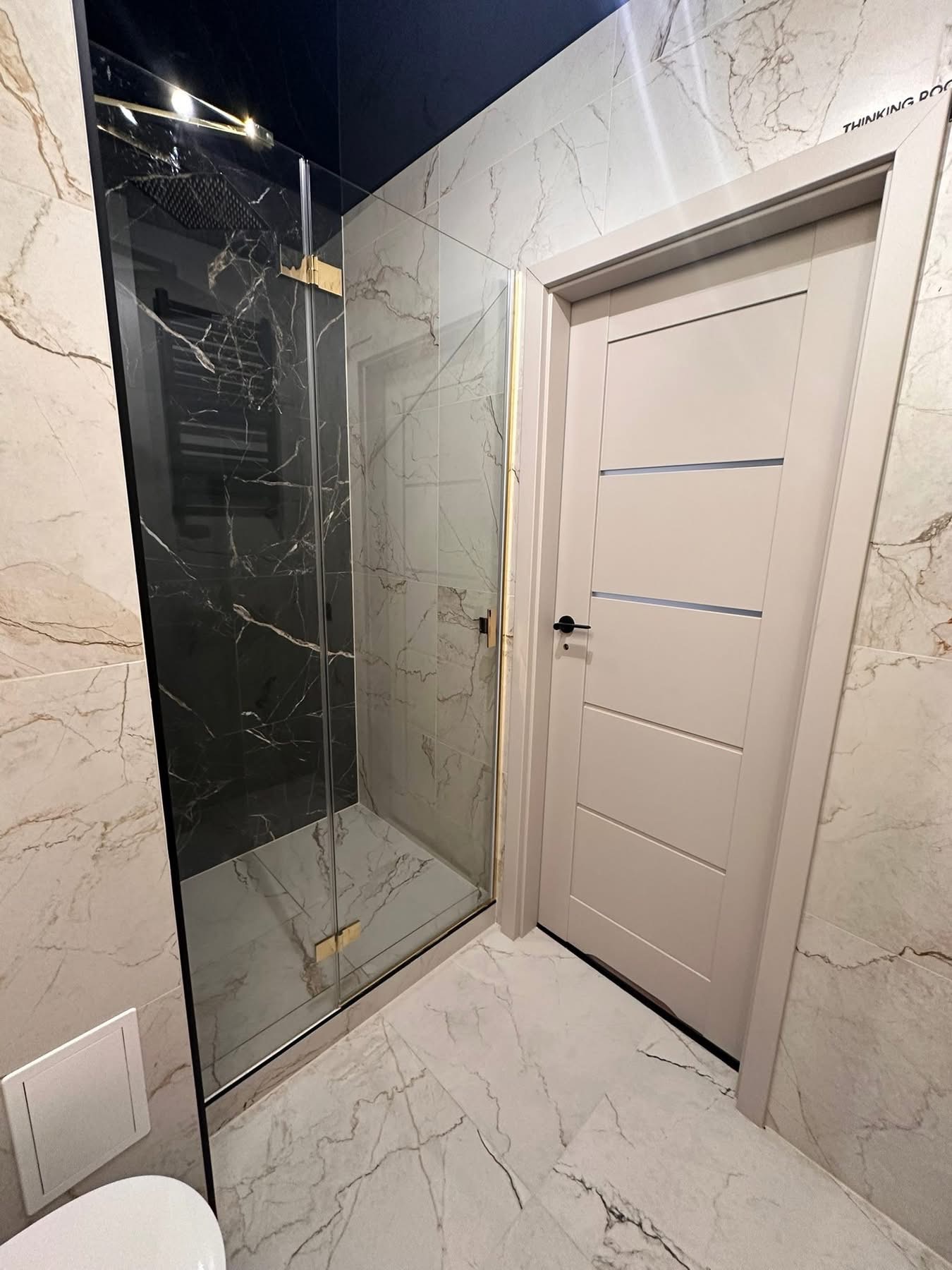 Nowoczesna łazienka z kabiną prysznicową typu walk-in z czarnym marmurem, jasnymi płytkami imitującymi marmur i białą, minimalistyczną stolarką drzwiową.