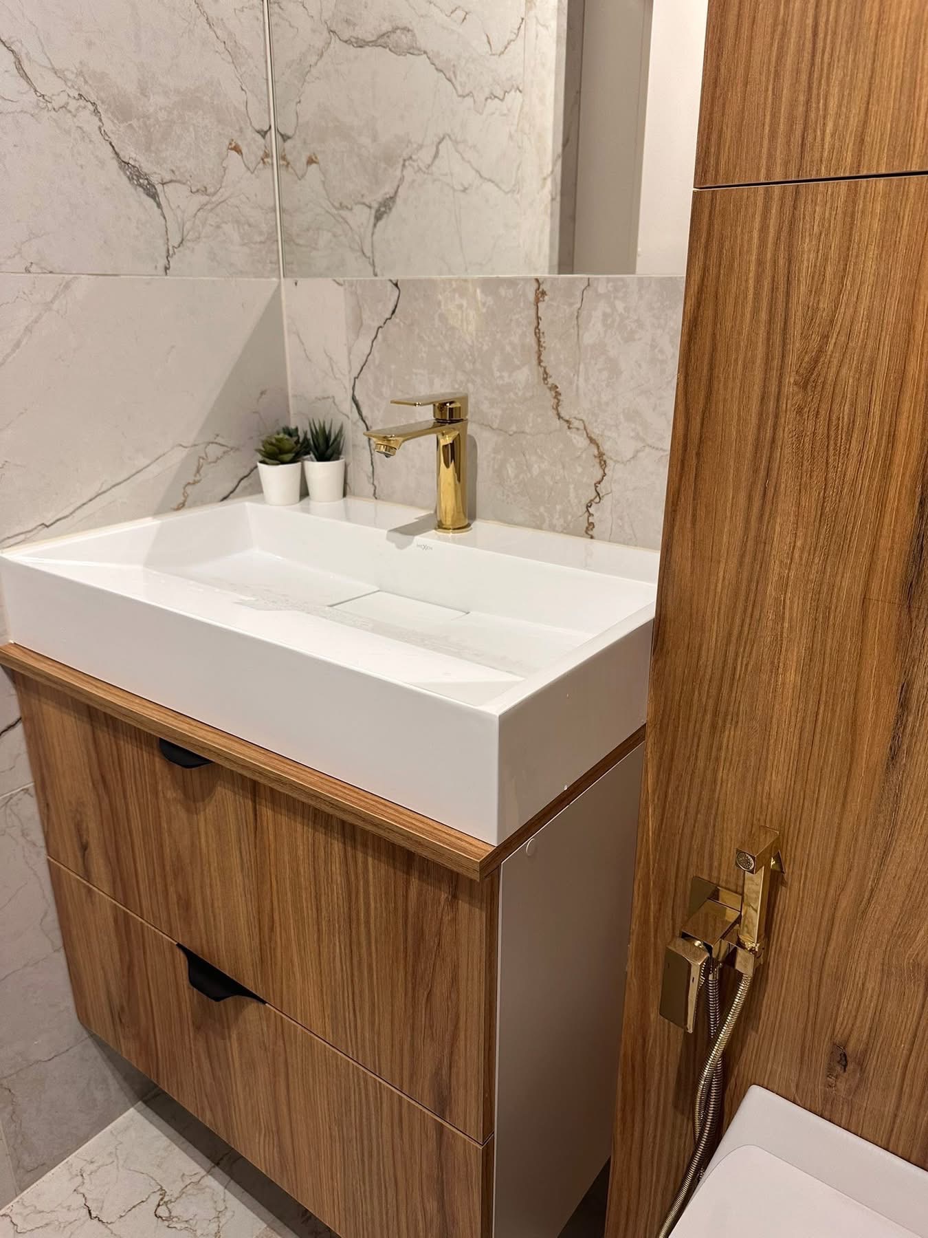 Nowoczesna łazienka z białą, prostokątną umywalką, złotą armaturą i drewnianą szafką z szufladami. Marmurowe płytki na ścianie i podłodze, roślinki w doniczkach.