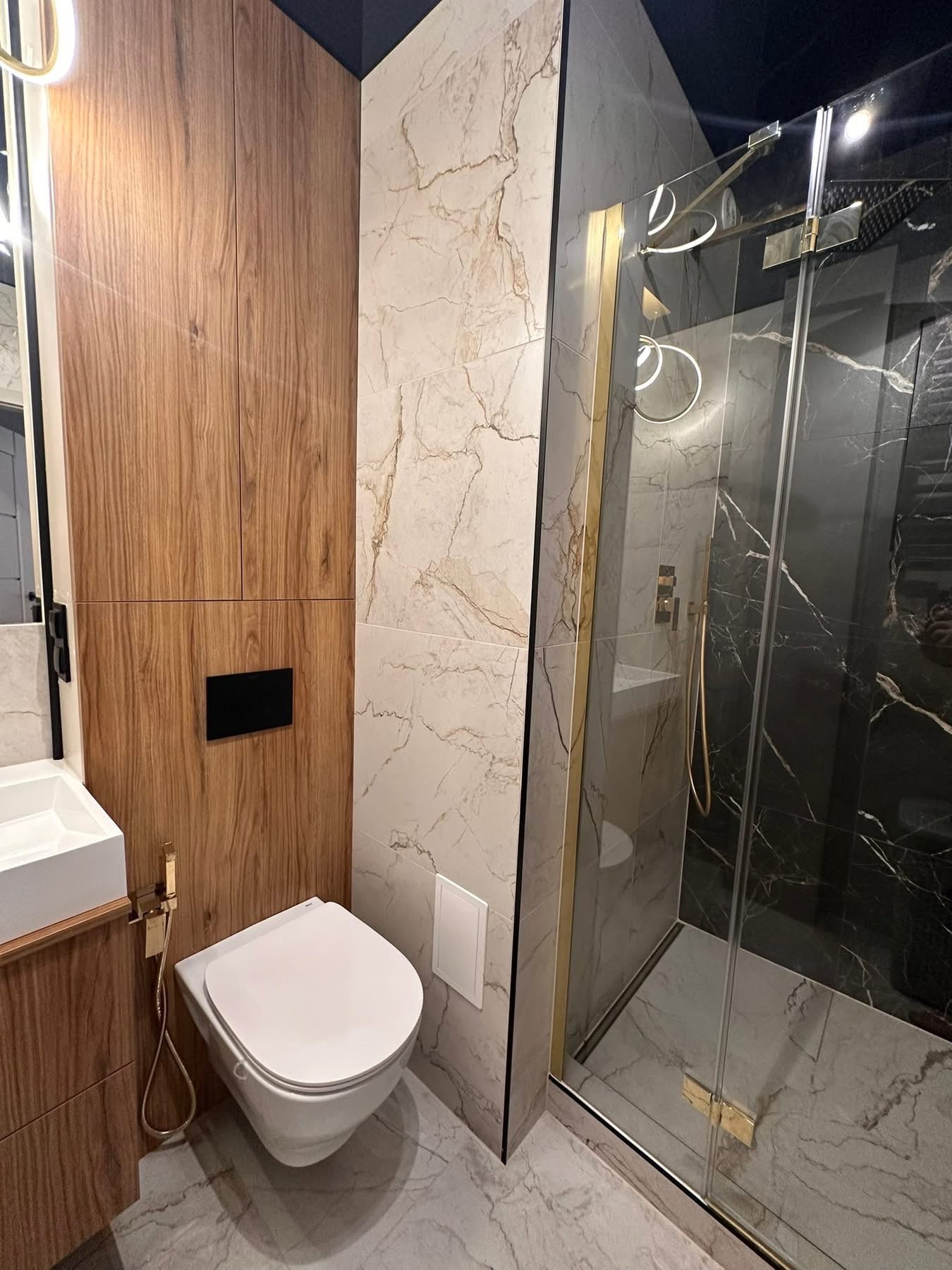 Nowoczesna łazienka z kabiną prysznicową typu walk-in, marmurowymi płytkami i drewnianą zabudową meblową. Uwagę zwracają złote akcenty i minimalistyczna armatura.